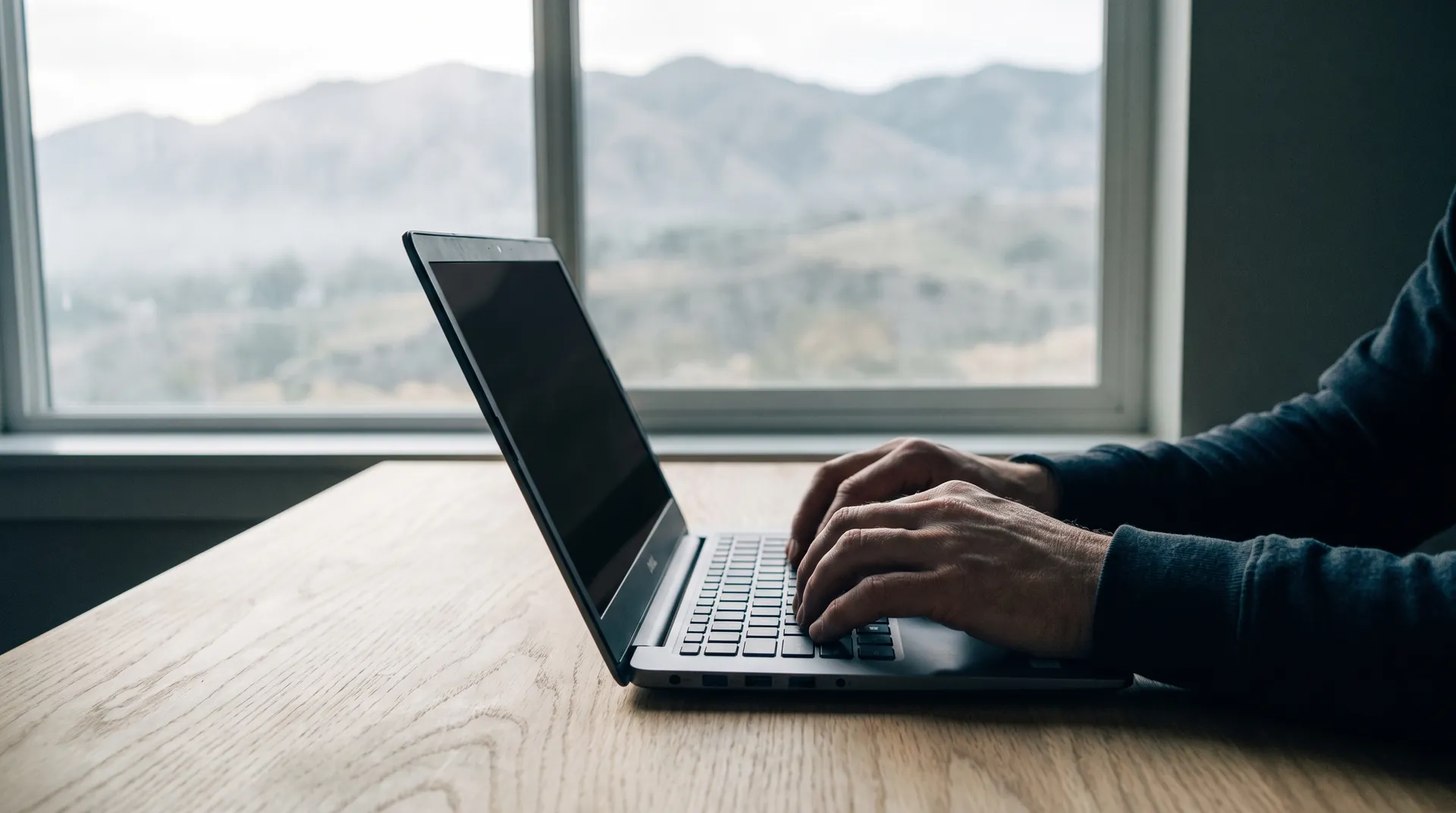 Hands typing on a laptop at a wooden table, foothills soft through the window behind