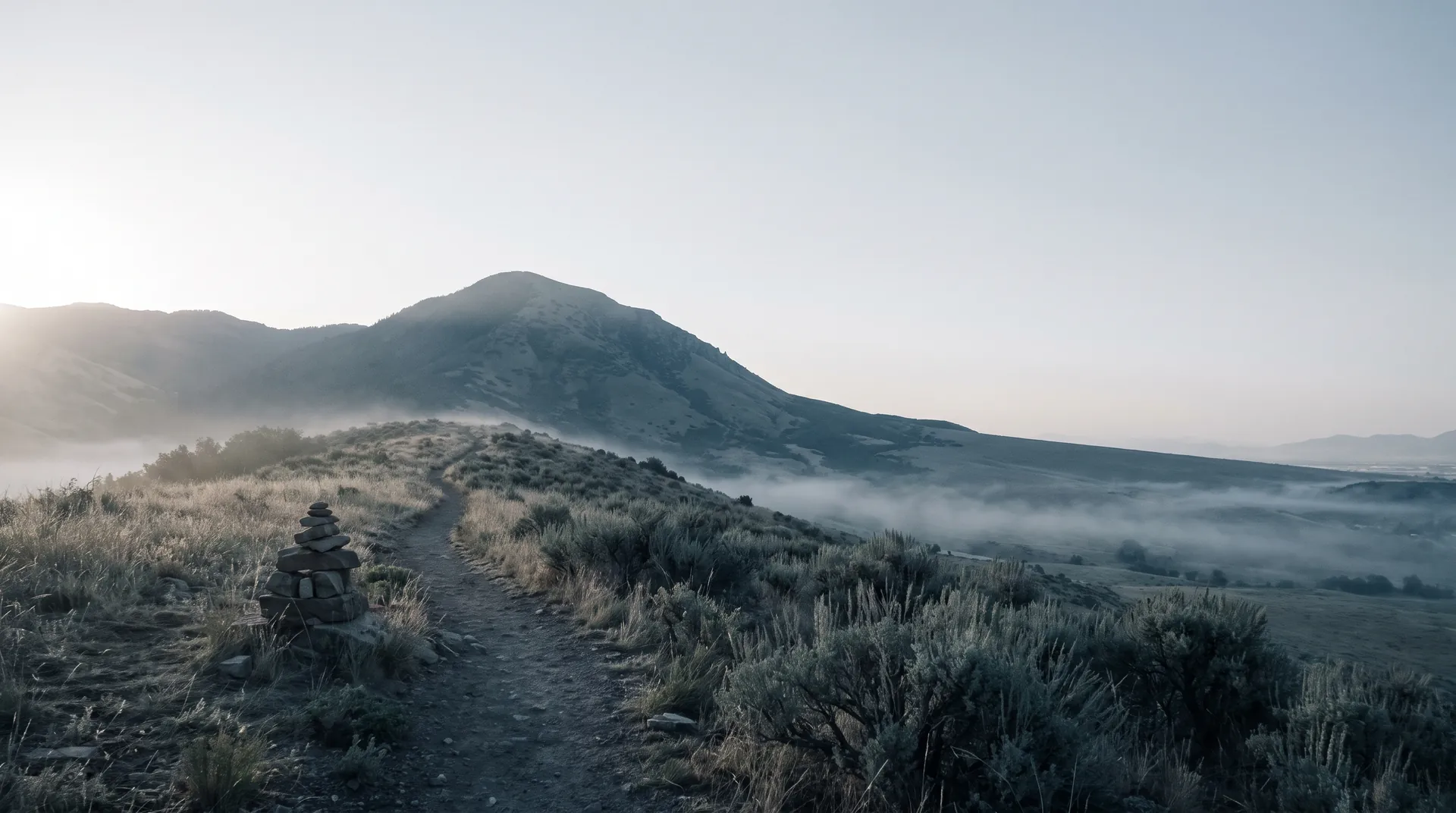 A stone trail cairn beside a dirt path on a sagebrush foothill ridge, mist lying in the valley below